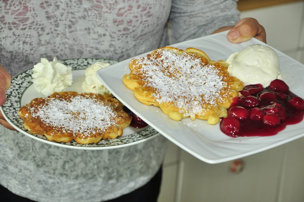 Zwei Teller mit frischen Waffeln, Puderzucker, Sahne, Eis und heißen Kirschen