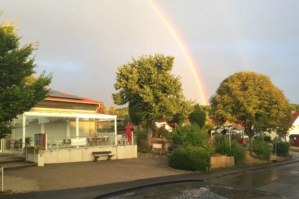 Der Dorfladen mit der Terrasse unter einem Regenbogen, davor der Platz und Bäume